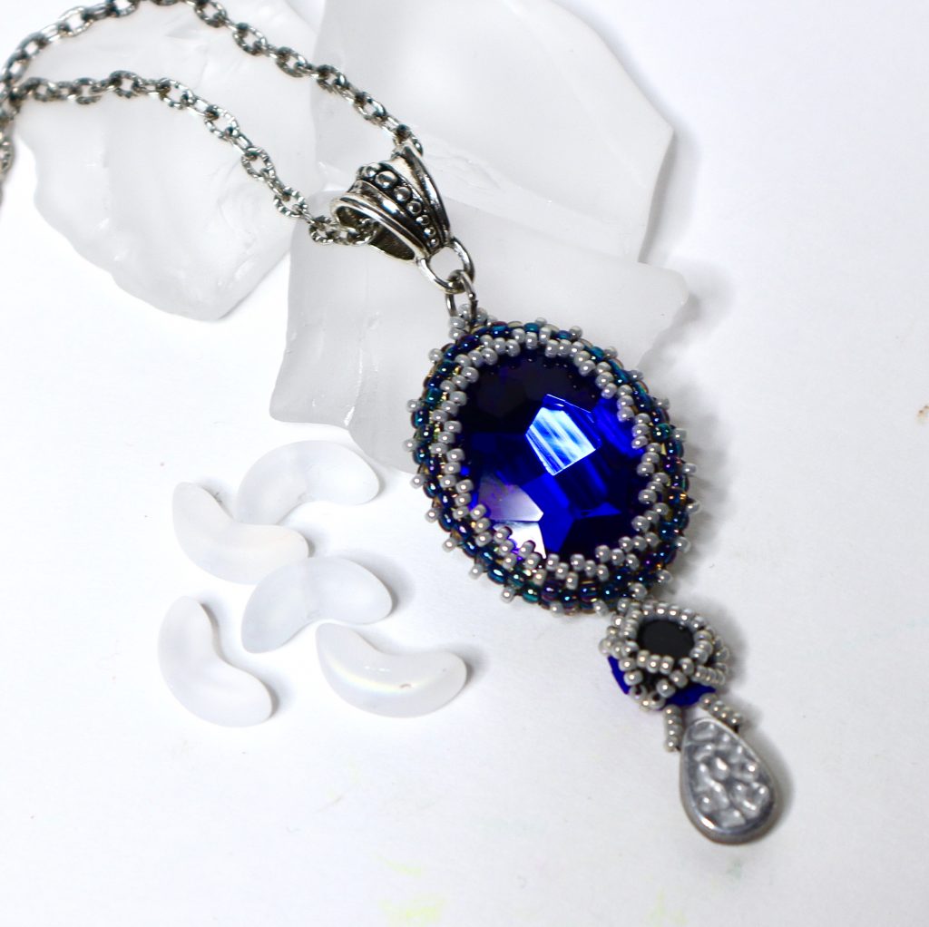 Deep sapphire blue oval crystal surrounded with gray and blue seed beads. 